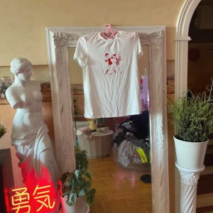 T-shirt No°2 - Vit t-shirt i bomull med korta ärmar och ett coolt rött tryck av två boxande tjejer på bröstet. En av figurerna har ett mönstrat plagg med cirklar och blommor. Perfekt för dig som gillar unika motiv och streetstyle.