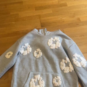 Grå hoodie från Denim Tears med blommor - Säljer en grå hoodie från Denim Tears med stora vita blommiga applikationer över hela tröjan. Hoodien har magficka och ribbade muddar. Perfekt för dig som gillar streetwear och vill sticka ut med en unik design.