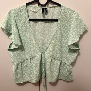 Mintgrön blommig blus från Vero Moda - Superfin mintgrön blus från Vero Moda med vitt blommönster. Blusen har korta ärmar, v-ringning och knytband i midjan. Perfekt till sommaren och lätt att matcha med jeans eller kjol.