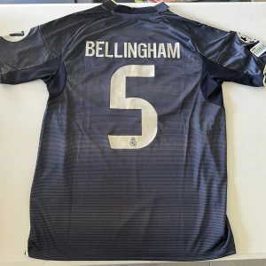 Real Madrid Bellingham bortatröja 23/24 - Säljer en mörkblå Real Madrid bortatröja för säsongen 23/24 med Bellingham och nummer 5 på ryggen. Tröjan har korta ärmar, tunna horisontella ränder, gula detaljer och Champions League-märken på ärmen. Tillverkad i lätt och ventilerande polyester med AEROREADY-teknologi. Replika, endast handtvätt gäller!