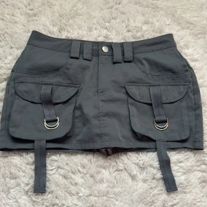 Grå utility minikjol från SHEIN XS - Grå utility-inspirerad minikjol från SHEIN med stora fickor framtill, dekorativa remmar och silverfärgade D-ringar. Kjolen har bälteshällor, knappstängning, bakfickor och inbyggda shorts. Perfekt för en edgy streetstyle-look. Använt någon enstaka gång, nypris 195kr.