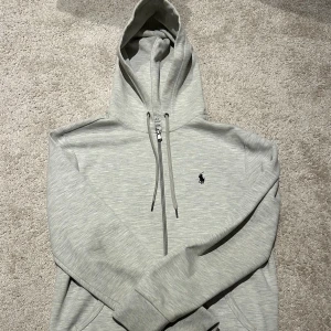 Ljusgrå hoodie från Polo Ralph Lauren - Snygg ljusgrå hoodie från Polo Ralph Lauren med dragkedja framtill, huva med snörning, klassisk broderad logga på bröstet och är i mycket bra skick. Tröjan har 2 fickor fram och är tillverkad i mjuk bomull och polyester. Perfekt för en chill och stilren look. 