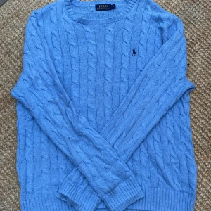 Ljusblå kabelstickad tröja Polo Ralph Lauren - Kabelstickad ljusblå tröja från Polo Ralph Lauren i 100% bomull. Klassisk rund halsringning, ribbade muddar och lång ärm. Ikoniska broderade loggan på bröstet. Perfekt för dig som gillar preppy och stilren look. Dock lite urtvättad tyvärr 