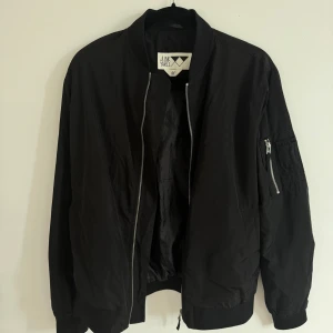 Svart bomberjacka från Junkyard - Svart bomberjacka från Junkyard i klassisk modell med ribbade muddar, silverfärgad dragkedja och ärmficka med zip. Jackan har en stilren look och är tillverkad i ett lätt syntetmaterial. Perfekt för dig som gillar streetstyle och enkel design.