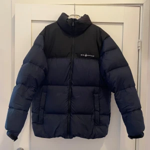 Svart och mörkblå Sail Racing dunjacka - Säljer en fet pufferjacka från Sail Racing i svart och mörkblått. Jackan är fylld med 600 fill power dun från Allied Feather & Down och har hög krage, dragkedja framtill och stora fickor. Perfekt för kalla dagar när du vill hålla stilen och värmen. Storleken är 176