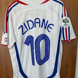 Frankrike Zidane VM 2006 - Säljer helt oanvänd  ikonisk fransk landslagströja från VM-finalen 2006 med Zidane 10 på ryggen. Tröjan är vit med blå och röda detaljer, broderat FFF-emblem och Adidas-logga. Korta ärmar, lätt och ventilerande material, klassisk passform. Perfekt för samlare eller fotbollsfans.
