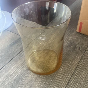 Fin vas från Holmegaard - Bilderna visar en Holmegaard-vas i brunt glas, med märkning 'HOLMEGAARD 1825'. Vasen har en elegant, modern design och ser ut att vara i gott skick utan synliga sprickor eller större slitage. Perfekt som dekorativ inredningsdetalj eller för blommor.