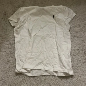 Vit t-shirt från Polo Ralph Lauren - Vit t-shirt från Polo Ralph Lauren med klassisk passform och korta ärmar. Liten svart broderad logga på bröstet. Tillverkad i mjuk bomull, perfekt för en clean och enkel stil.