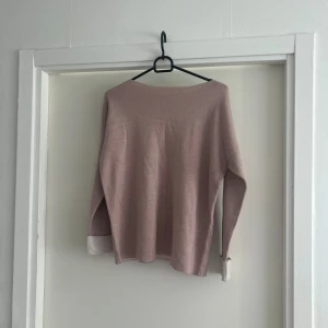 Ljusrosa stickad tröja med vita muddar - Mysig ljusrosa stickad tröja med långa ärmar och vita muddar. Tröjan har en enkel, rak passform och är perfekt för dig som gillar en clean och soft look. Materialet är mjukt och känns skönt mot huden. Snygg att matcha med jeans eller kjol.