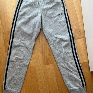 Grå Adidas mjukisbyxor med svarta ränder - Grå mjukisbyxor från Adidas med klassiska svarta ränder längs sidorna och Adidas-logga på låret. Byxorna har resår och snörning i midjan samt muddar vid bensluten. Tillverkade i mjukt material som är skönt mot huden.