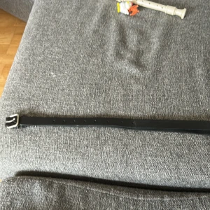 Svart klassiskt skärp i skinn - Snyggt svart bälte i äkta skinn med enkel, fyrkantig metallspänne i silverfärg. Bältet har en stilren och tidlös design med flera hål för justerbar passform. Perfekt accessoar för att lyfta vilken outfit som helst.