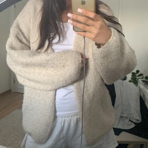 Beige stickad kofta från Lindex - Mysig och oversized beige stickad kofta från Lindex. Koftan har lång ärm, öppen front och är tillverkad i ett fluffigt, grovstickat material som känns supergosigt. Perfekt att slänga på sig när det är kyligt ute.