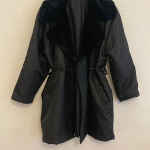 Svart parkajacka med fuskpäls - Säljer en svart jacka med stor krage i fuskpäls. Jackan har långa ärmar, två fickor framtill. Bälte saknas då den är inköpt på second hand, men öglor finns.  Perfekt för lite kyliga dagar och har en klassisk, stilren look.