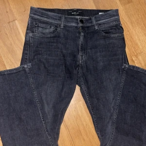 Replay svarta jeans Thad  - Snygga svarta jeans från Replay med klassisk femficksdesign och raka ben. Jeansen har diskreta slitningar och tydliga sömmar, samt Replay-logga på bakfickan. Perfekta för dig som gillar en stilren look med lite edge.