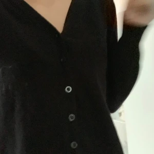 Svart kofta med knappar - Svart kofta med v-ringning och knäppning framtill. Långärmad och i mjukt material, perfekt att slänga över en topp eller bära stängd. Enkel och stilren design som funkar till det mesta.