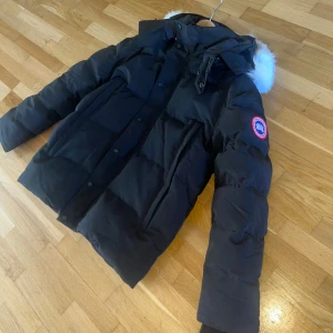 Svart Canada Goose Wyndham Parka XL - Svart Wyndham Parka från Canada Goose i storlek XL. Jackan har en puffer-design med dunfyllning, huva med äkta päls och klassisk logopatch på ärmen. Flera fickor framtill, dragkedja och knappar. Perfekt för kalla vinterdagar. Säljes pga fel storlek, kan även bytas mot Goose eller annan vinter jacka i M/L