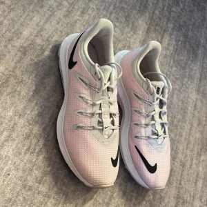 Nike sneakers i ljusrosa och vitt - Snygga Nike sneakers i ljusrosa mesh med vita och grå detaljer samt svarta Nike-loggor på sidan och framtill. Skorna har snörning och en sportig, modern design med platt sula. Perfekta för dig som gillar stilrena och bekväma sneakers.