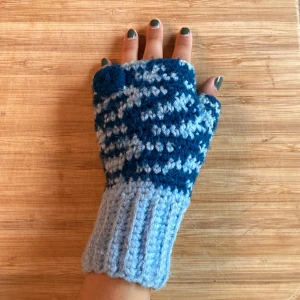 Blå virkade fingerlösa vantar - Ett par handgjorda fingerlösa virkade vantar med coolt mönster i ljusblått och mörkblått. Perfekta när du vill hålla händerna varma men ändå kunna använda mobilen. Ribbstickad mudd för extra komfort och snygg passform. Använda av mig tidigare men i bra skick
