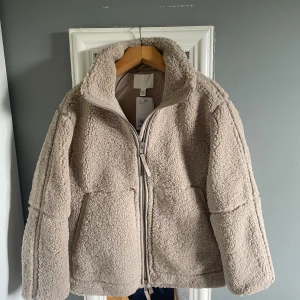 Beige teddyjacka från H&M XXS - Supermjuk och fluffig teddyjacka i beige från H&M. Jackan har hög krage, dragkedja framtill och två stora fickor. Perfekt för kyliga dagar och ger en riktigt cozy vibe. Materialet är polyester och jackan har en loose passform. Den är helt ny och aldrig använd.