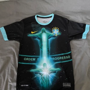 Brazil x jesus t-shirt - Snygg svart Brasilien fotbollströja från Nike med Neymar Jr och nummer 10 på ryggen. Framsidan har ett stort tryck av Kristusstatyn och texten 'Ordem e Progresso'. Tröjan har ljusblå detaljer vid krage och ärmslut.