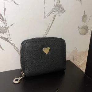Svart plånbok med silverfärgat hjärta - Snygg svart plånbok med silverfärgat hjärta på framsidan och praktisk dragkedja runtom. Insidan har flera fack för kort och mynt, samt ett extra fack med dragkedja. Perfekt storlek för att ha med sig överallt.