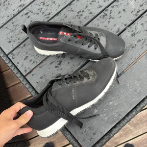 Svarta sneakers från Prada - Svarta sneakers från Prada med vit sula och klassisk röd logga på plösen. Skorna har snörning och är tillverkade i syntetmaterial med detaljer i skinn. Snygg och stilren design som passar till många outfits. Använda 1 gång men lite smutisga, inget som inte går bort på en tvätt nypris 4300