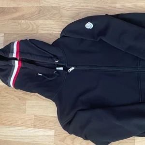 Svart Moncler hoodie med röd/vit detalj - Svart hoodie från Moncler med dragkedja, huva och snörning. Ärmslut och huva har snygga rödvita ränder. Klassisk Moncler-logga på ena ärmen. Perfekt för dig som vill ha en stilren och exklusiv look.