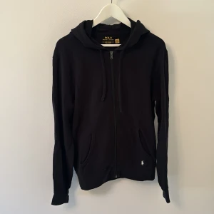 Polo Ralph Lauren hoodie  - Svart hoodie från Polo Ralph Lauren med dragkedja framtill, huva med snörning och två fickor. Klassisk design med liten broderad logga vid fickan. Perfekt för en chill och stilren look.