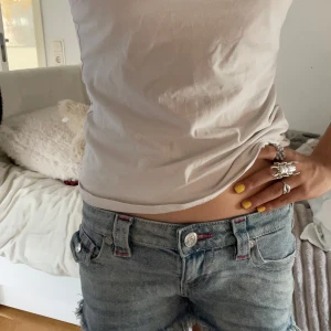 True Religion lågmidjade jeansshorts - Säljer ett par blå jeansshorts från True Religion, modell Joey Cut-Off. De är lågmidjade med råa, fransiga kanter och har röda detaljer vid sömmarna och fickorna. Klassiska bakfickor med broderad logga och silverfärgade knappar. Perfekta för sommaren! Andvänd max 3 gånger  Skulle gärna träffa🫶🏼💞
