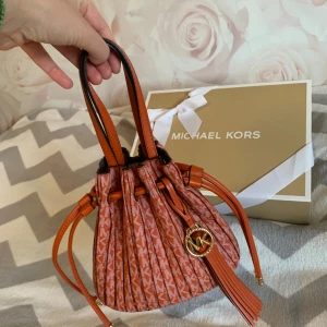 Michael Kors orange handväska med snörning - Superfin orange handväska från Michael Kors med MK-logga och tofsdetalj i guld. Väskan har snörning upptill, två handtag och avtagbar axelrem. Mönstret består av små vita MK-bokstäver över hela väskan. Perfekt storlek för det viktigaste och riktigt snygg design.