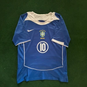 Blå Brasil Ronaldinho Nike fotbollströja - Säljer en blå Brasilien fotbollströja från Nike med vita detaljer och nummer 10 på både fram- och baksidan. På ryggen står Ronaldinho. Tröjan har korta ärmar och är gjord i lätt, ventilerande material. Perfekt för dig som älskar fotboll och ikoniska spelare.