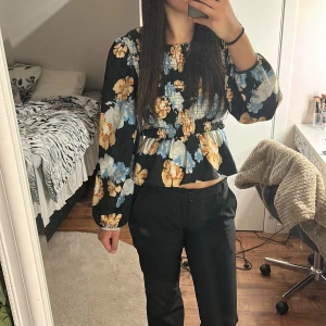 Blommig blus från ONLY Studio - Supersöt blommig blus från ONLY Studio med smockad överdel och volang nertill. Svart botten med stora blommor i blått, beige och vitt. Långa puffärmar och rund halsringning. Perfekt för dig som gillar färg och mönster!