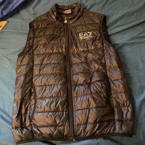 Svart Emporio Armani EA7 väst - Snygg svart dunväst från Emporio Armani (EA7) med quiltad design och silverfärgat logotryck på bröstet. Västen har ståkrage, dragkedja framtill och två sidofickor. Perfekt att slänga över en hoodie för en chill look. Nypris 1950kr från Zalando. Pris kan diskuteras🙂