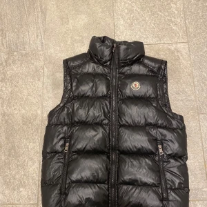 Moncler väst svart med/utan luva - Snygg svart dunväst från Moncler med avtagbar huva och två dragkedjefickor framtill. Västen har quiltad design och Moncler-logga på bröstet. Perfekt för lager-på-lager och riktigt skön att bära under kyliga dagar. Priset kan diskuteras under snabb affär 