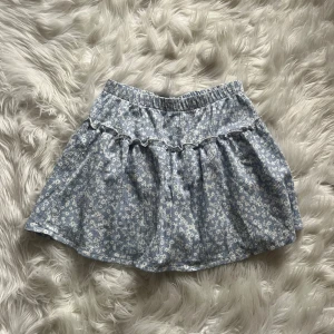 Blommig blå kortkjol från Zara - Supersöt blå kortkjol från Zara med vitt blommigt mönster och volangdetalj. Kjolen har en skön resår i midjan och är gjord i mjuk bomull, perfekt för vår och sommar. Passformen är normal och kjolen är lätt att matcha med en enkel topp.