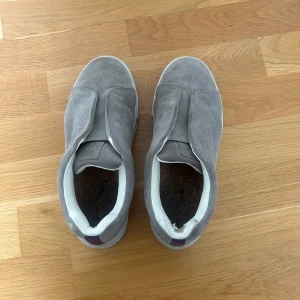 Grå mockasneakers med vit sula - Snygga grå sneakers i mocka med vit platt sula och elastisk detalj över vristen för enkel påtagning. Skorna har en stilren och minimalistisk look. Finns en lite defekt på insidan vid höger häl, syns dock inte när man har skorna på sig 
