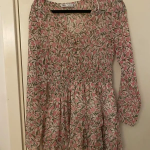 Blommig kortklänning från Zara - Supersöt kortklänning från Zara med rosa och grönt blommönster på ljus botten. Klänningen har v-ringning, knappar framtill, smock i midjan och långa ärmar. Perfekt för vår och sommar, flowig och lätt i materialet.