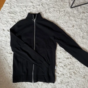 Svart kofta med dragkedja från Selected Homme - Säljer en svart kofta från Selected Homme i storlek S. Tröjan har hel dragkedja framtill, hög krage och diskret strukturmönster på bröst och ärmar.