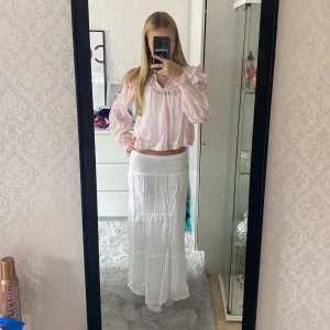 Vit långkjol i bomull - Luftig och flowy vit långkjol med smock i midjan och flera volanger. Kjolen är tillverkad i tunn bomull och har en avslappnad, bohemisk vibe. Perfekt för varma dagar och enkel att styla med crop tops eller blusar.