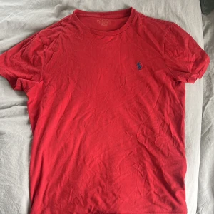 Röd t-shirt från Polo Ralph Lauren - Snygg röd t-shirt från Polo Ralph Lauren med klassisk blå broderad logga på bröstet. Modellen är slim fit och har rund hals samt korta ärmar. Tillverkad i mjuk bomull som är skön mot huden. Perfekt för dig som gillar stilrena och ikoniska plagg.