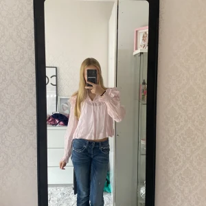 Ljusrosa blus med volangdetaljer - Säljer en ljusrosa blus med volangdetaljer på axlarna och ärmarna. Blusen har långärmade, lätt transparenta ärmar med spetsinslag och en lös, croppad passform. Perfekt för dig som gillar romantisk och feminin stil.