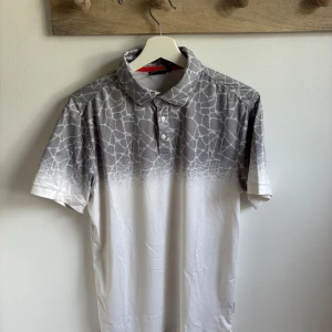 Gråvit mönstrad piké från Func Factory - Snygg piké från Func Factory i storlek M med coolt grått och vitt mönster upptill som liknar stenar. T-shirten har krage, knappar och är tillverkad i 100% polyester vilket gör den lätt och skön. Perfekt för dig som gillar sportig och modern stil.