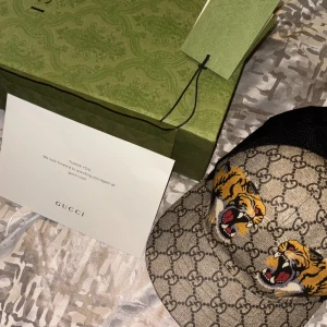 Gucci keps med tigertryck och GG-mönster - Snygg keps från Gucci med klassiskt GG-monogram i beige och brunt, prydd med två broderade tigrar framtill. Baksidan är i svart mesh för extra ventilation och insidan har den ikoniska gröna och röda Gucci-randen. Perfekt för dig som vill sticka ut med lyxig streetstyle.