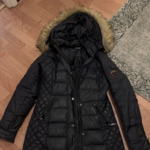 Svart jacka, Rockandblue - Snygg svart jacka från Rockandblue med quiltade partier och fuskpäls på huvan. Jackan har dragkedjor på fickorna, emblem på ärmen och en klassisk pufferstil. Perfekt för kalla dagar när du vill hålla stilen.