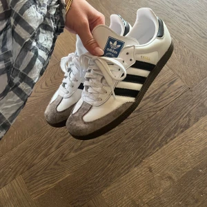 Adidas Samba vita sneakers - Klassiska Adidas Samba sneakers i vitt skinn med svarta ränder och grå mockadetaljer framtill. Skorna har snörning, platt sula och blå logga på plösen. Perfekta för en sportig och trendig look.