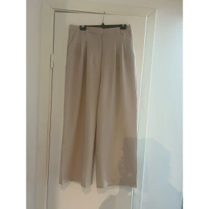 Beiga wide kostymbyxor med pressveck - Snygga beiga kostymbyxor med vida ben och pressveck framtill. Byxorna har hög midja, hällor för bälte och fickor i sidorna. Perfekta för en clean och stilren look. Materialet känns mjukt och har ett fint fall.