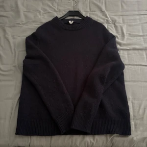 Mörkblå stickad tröja i storlek L, arket - Säljer en enkel och stilren mörkblå stickad tröja i storlek L från Arket. Tröjan har rund hals med bred ribbad kant och långa ärmar. Perfekt för dig som gillar klassiska och bekväma plagg med lite loose passform. Materialet är mjukt och värmande, perfekt för kyliga dagar.