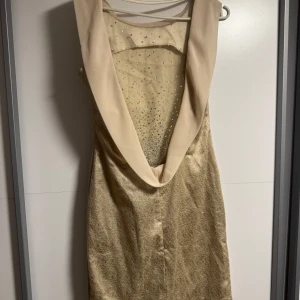 Champagnefärgad kortklänning med paljetter - Säljer en elegant kortklänning i champagnefärg med glittriga paljettdetaljer framtill. Klänningen har en djup v-ringning med bred krage och är ärmlös. Materialet har en lyxig satinliknande finish som ger en exklusiv känsla. Perfekt för dig som vill sticka ut.