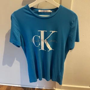 Blå Calvin Klein Jeans t-shirt - Snygg blå t-shirt från Calvin Klein Jeans med stort vitt CK-tryck på bröstet. Klassisk rund halsringning och korta ärmar. Perfekt för dig som gillar stilrena och trendiga plagg med tydlig logga. Tillverkad i mjuk bomull för skön känsla.