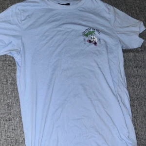 Ljusblå t-shirt från SMOG XS - Ljusblå t-shirt från SMOG i bomull med tryck fram och stort tryck på ryggen med texten 'Holiday in Hell' och en dödskalle med palmer. Regular fit och kortärmad, perfekt för chill dagar. Snygg och unik design för dig som vill sticka ut.